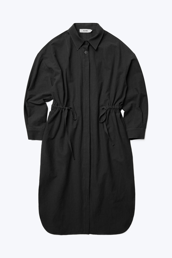 The front of the Filter Dress Black, a classic style with a button through partly concealed placket and built in collar, long sleeves, in seam side pockets and rounded scoop hemline. Featuring waist ties to adjust the waistline to your liking.