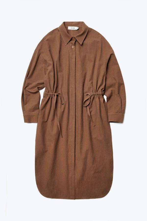 The front of the Filter Dress Brown, a classic style with a button through partly concealed placket and built in collar, long sleeves, in seam side pockets and rounded scoop hemline. Featuring waist ties to adjust the waistline to your liking. Made from 100% cotton.