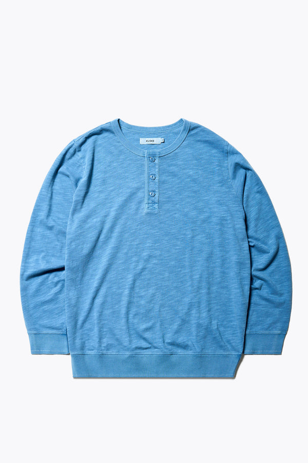 The Asperse Henley in Blue, featuring a three-button placket, ribbed cuffs and hem, and a garment pigment-dyed finish. Available in 5 sizes.