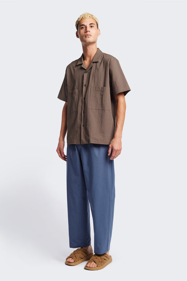 The Boulder Pleat Pants in Blue, crafted from cotton herringbone twill, featuring side slant pockets, back patch pockets, and an elastic waistband with internal pull. Available in 5 sizes.