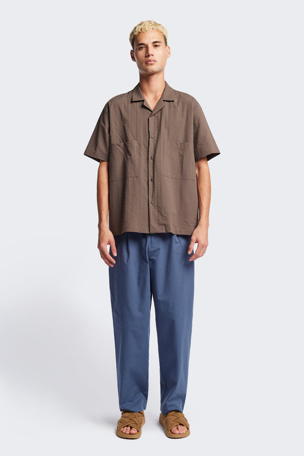 The Boulder Pleat Pants in Blue, crafted from cotton herringbone twill, featuring side slant pockets, back patch pockets, and an elastic waistband with internal pull. Available in 5 sizes.