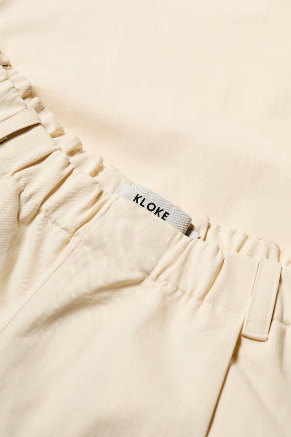 Close up details of The Boulder Pleat Pants in Chalk Beige, crafted from cotton herringbone twill, featuring side slant pockets, back patch pockets, and an elastic waistband with internal pull. Available in 5 sizes.