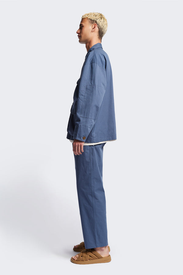 Side profile of The Pressure Jacket in Blue, a relaxed boxy long sleeve jacket in washed cotton herringbone with 3D patch pockets and hidden zip openings. Available in 5 sizes.