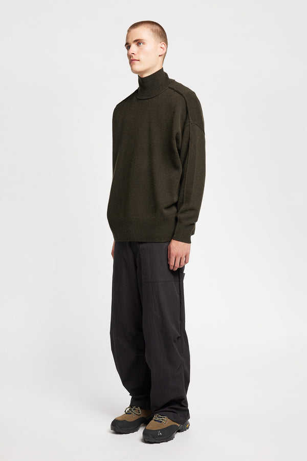 Front angle of Model wearing mock neck merino wool fisherman sweater with exposed seams in Darkest Green.
