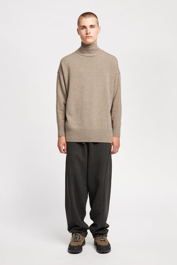 Front of Model wearing mock neck merino wool fisherman sweater with exposed seams in Taupe.