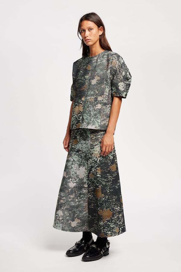 Front view model in the Optic Skirt a midi-length A-line skirt in textured jacquard green print fabric with a forest-inspired pattern, designed by Kloke