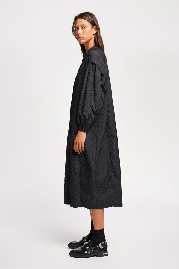 Side of Model wearing the Parapet Dress in Jet Black. A lightweight seersucker nylon, showing band collar and adjustable front waist tie.