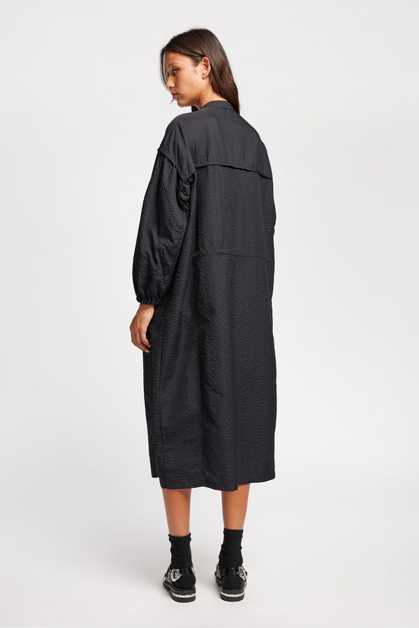 Back of Model wearing the Parapet Dress in Jet Black. A lightweight seersucker nylon, showing band collar and adjustable front waist tie.