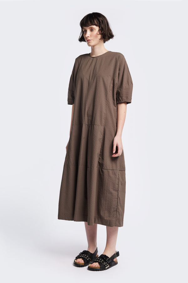The Scoria Dress in brown, a long-panelled cotton seersucker dress with voluminous sleeves, structured panel details, and a fuller skirt, perfect for an effortless summer look. Available in 5 sizes.