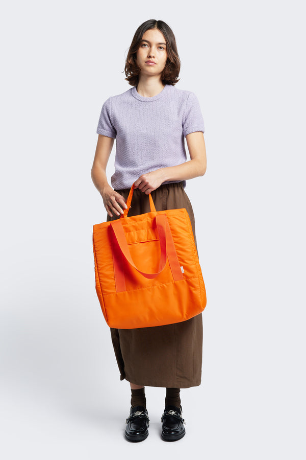 Model holding the short straps on the Influx Padded Bag Orange, a practical tote with four compartments including two snap-closing pockets. 