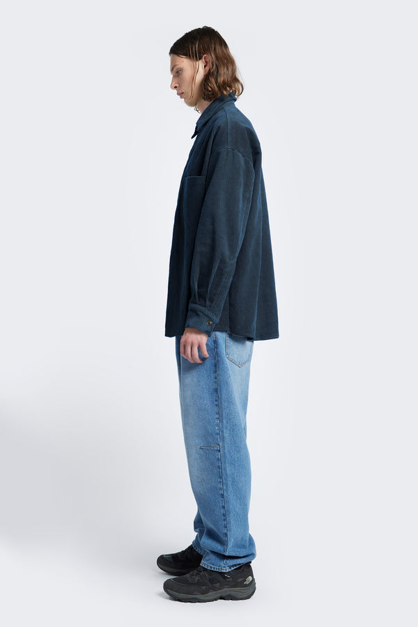 Side profile of the Corner Cord Shirt in Petrol Blue, a long sleeve shirt with rounded hemline and made from thick wale corduroy. 