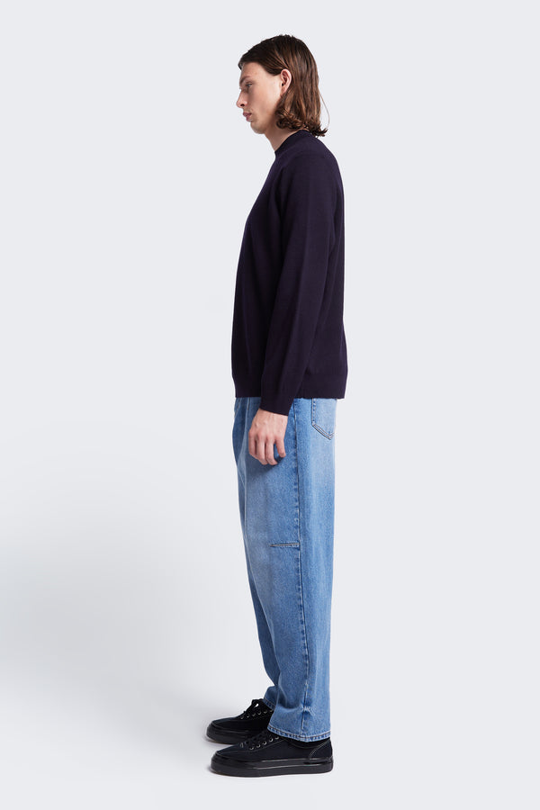 Model stands side on wearing the Dose  Merino Sweater Ink, knitted in a fine Merino wool in a milano knit construction, this mock-neck style is finished with a 1x1 rib hemline and cuffs. 
