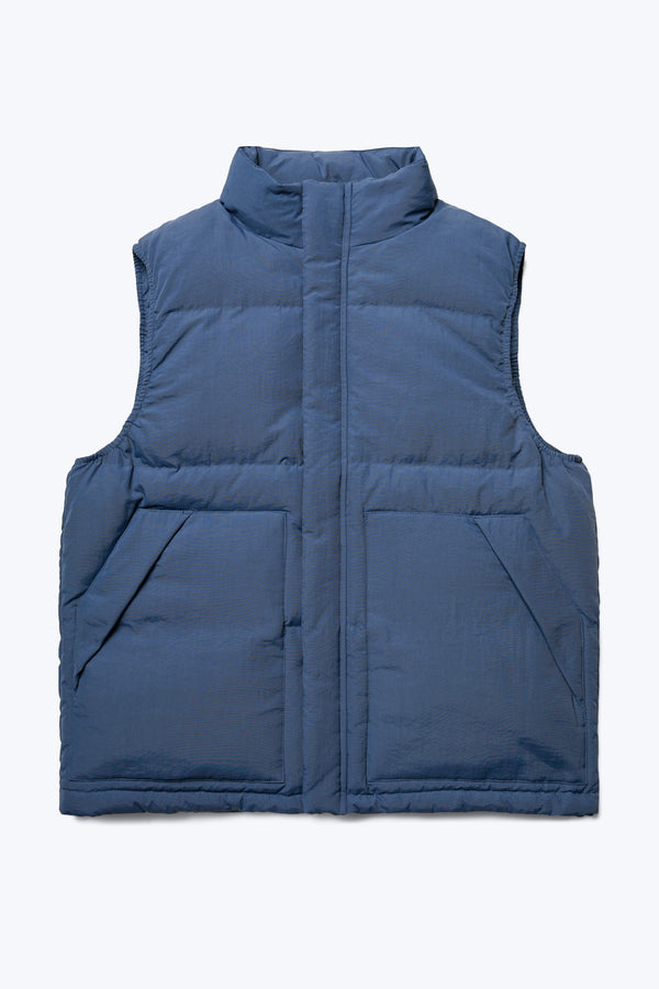 The front of the Reset Quilted Puffer Vest Blue, with a high neck, centre zip and stud closure, internal and side pockets and adjustable toggled elastic waist. Made with 100% post-consumer down fill. Available in five sizes. 