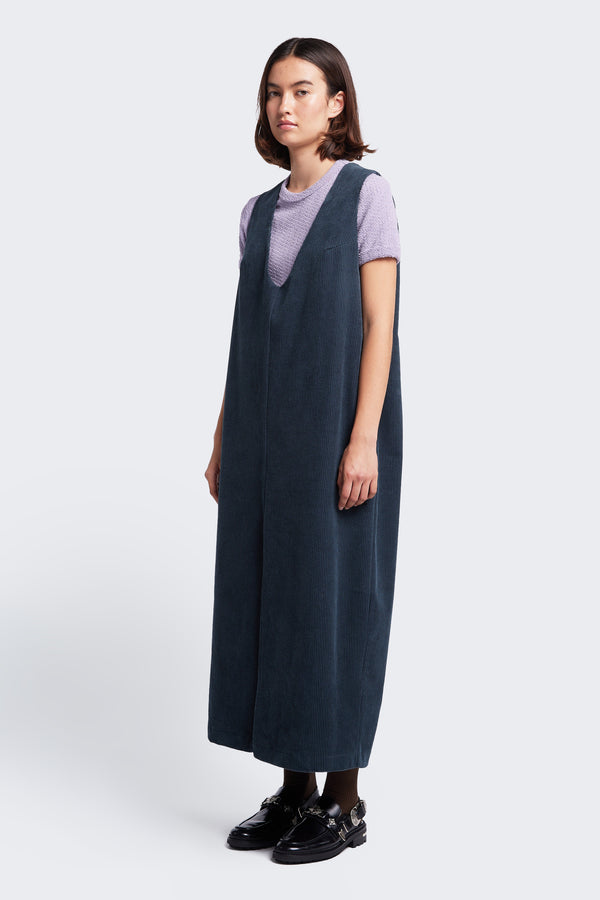 The Arc Dress, a full-length pinafore made from thick-wale corduroy in Petrol Blue featuring a deep V neckline and front centre split. Styled with a purple tee. 