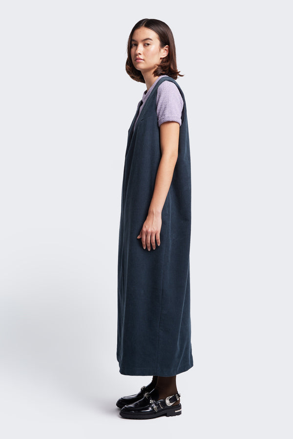 The Arc Dress Petrol Blue, a full length pinafore made from a thick wale corduroy. Styled with the Exodus Tee Lilac.