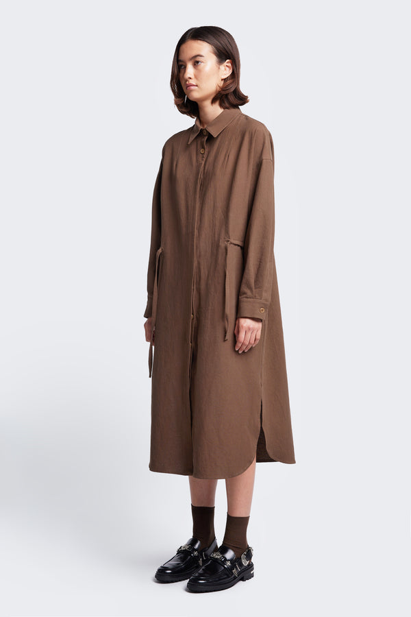 The Filter Dress Brown crafted in 100% cotton. Featuring waist ties to adjust the shape to your liking. The dress has a centre button through partly concealed placket, collar, long sleeves, in seam side pockets and rounded scoop hemline with side split.