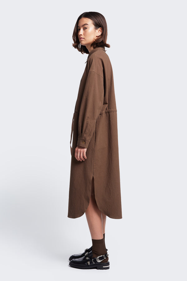 Side profile of the Filter Dress with long sleeves, rounded hemline with side split and waist ties to adjust the shape to your liking. Made from 100% cotton in Brown. 