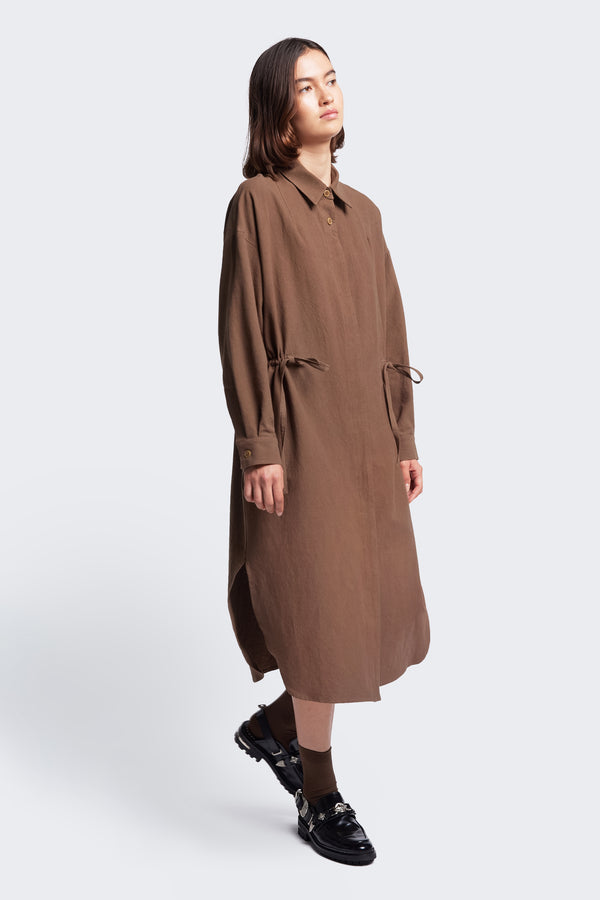 The Filter Dress crafted in 100% cotton in Brown. Featuring waist ties to adjust the waistline and shape to your liking. The dress has a centre button through partly concealed placket, built in collar, long sleeves and rounded scoop hemline with side split.