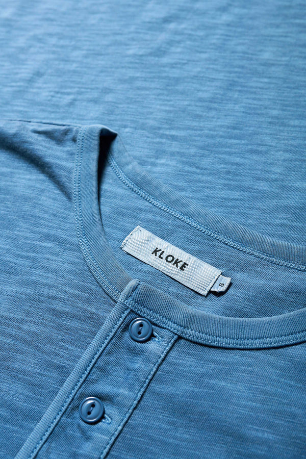 Close up details of The Asperse Henley in Blue, featuring a three-button placket, ribbed cuffs and hem, and a garment pigment-dyed finish. Available in 5 sizes. 