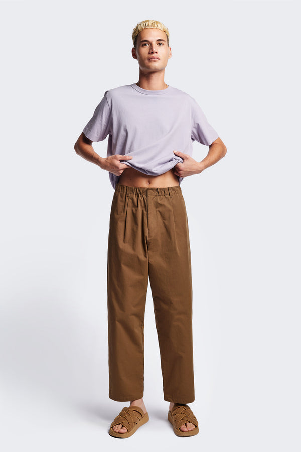 The Boulder Pleat Pants in Brown, crafted from cotton herringbone twill, featuring side slant pockets, back patch pockets, and an elastic waistband with internal pull. Available in 5 sizes. 