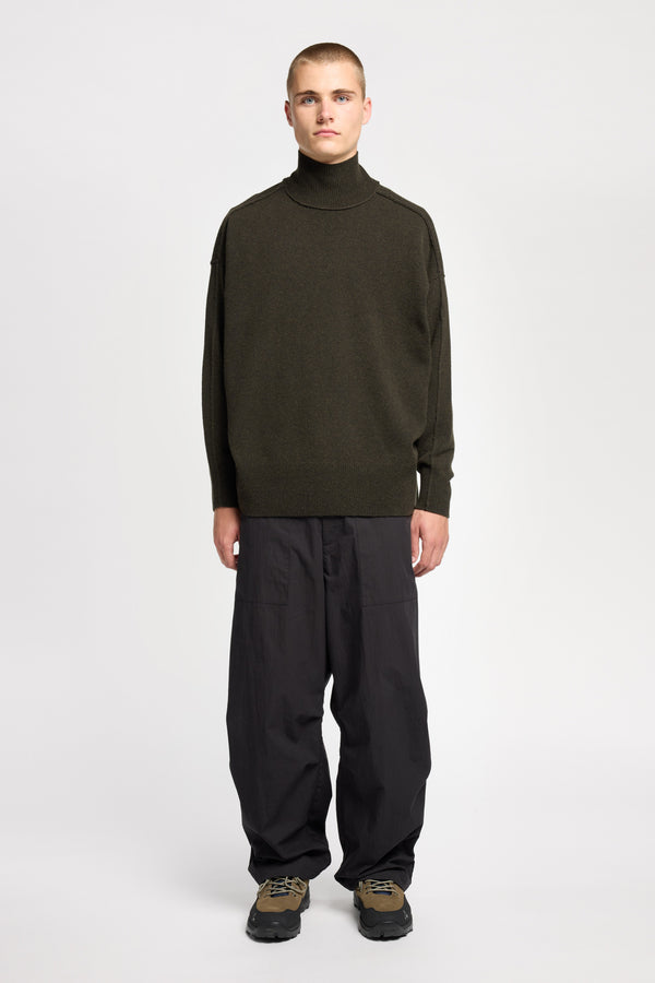Front of Model wearing mock neck merino wool fisherman sweater with exposed seams in Darkest Green. 