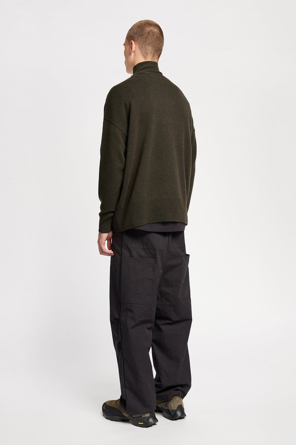 Back angle of Model wearing mock neck merino wool fisherman sweater with exposed seams in Darkest Green. 