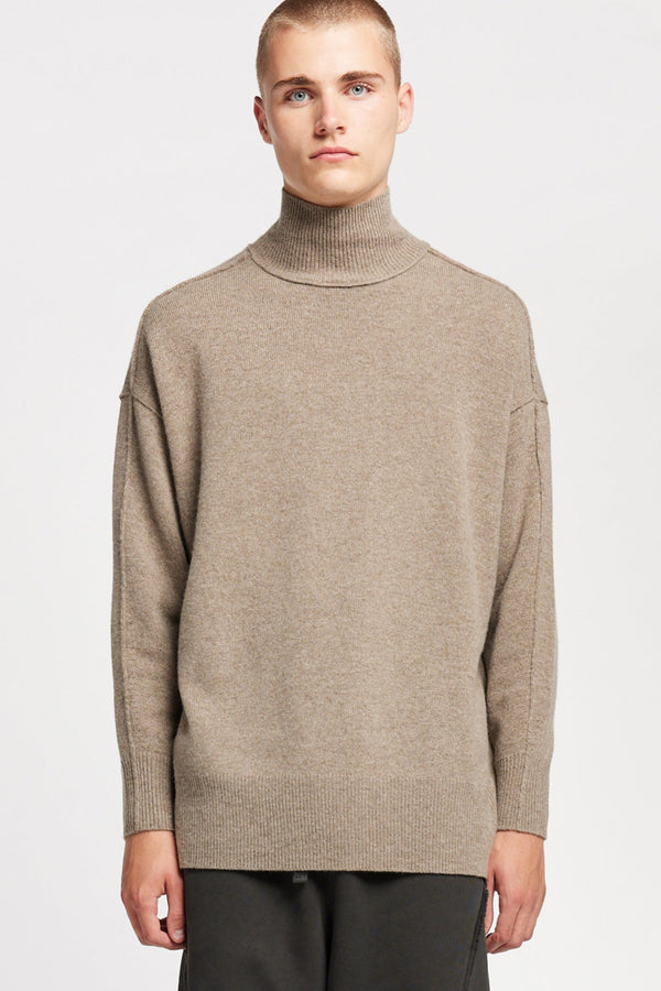 Front of Model wearing mock neck merino wool fisherman sweater with exposed seams in Taupe. 