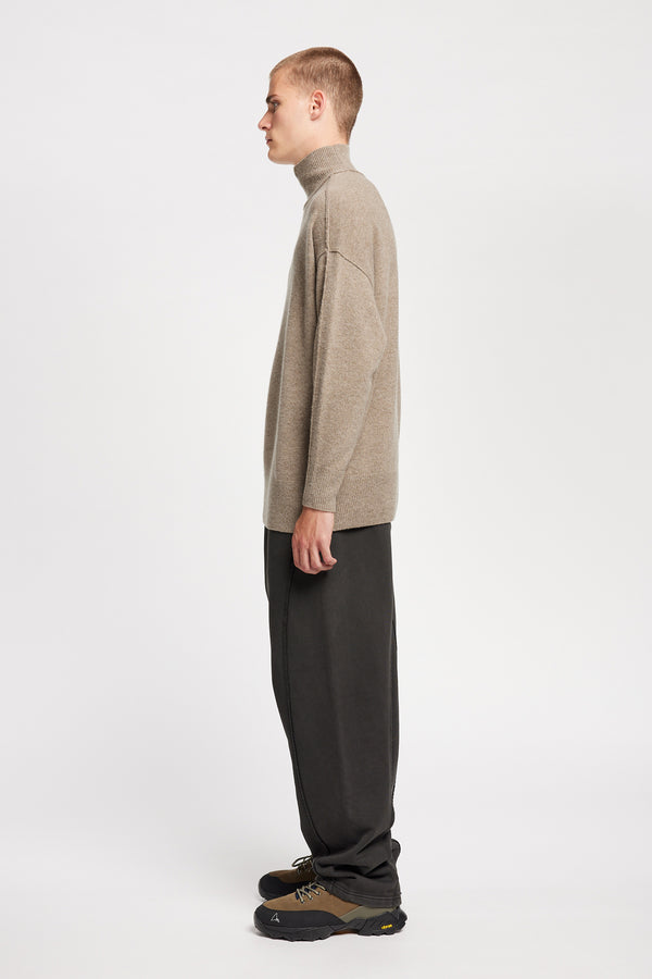 Side  of Model wearing mock neck merino wool fisherman sweater with exposed seams in Taupe. 