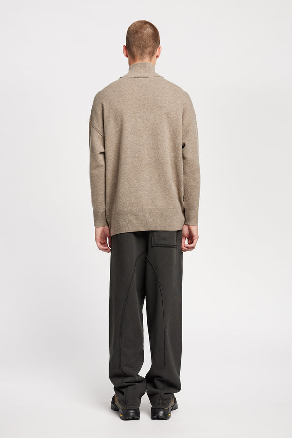 Back of Model wearing mock neck merino wool fisherman sweater with exposed seams in Taupe. 
