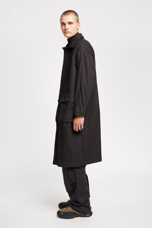 Side view of model wearing the venture trench coat in jet black. An oversized fit in cotton-nylon with structured collar and flap pockets