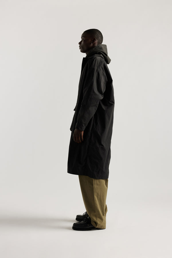 Side view of male model wearing the venture trench coat in jet black. An oversized fit in cotton-nylon with structured collar and flap pockets