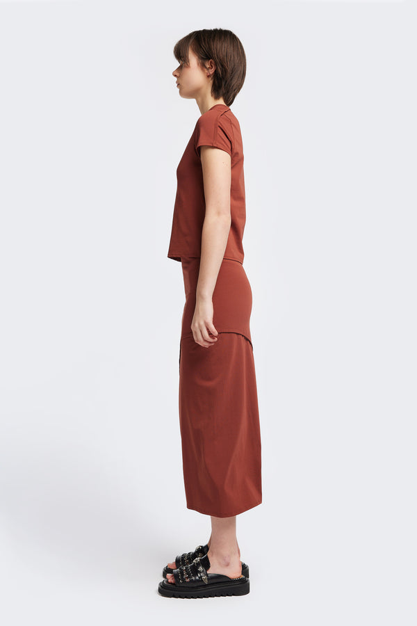 Side view of The Cumulus Skirt in brick red. A Slim, mid-calf dress in a cotton blend with stretch, featuring a curved bound seam for a flattering silhouette.