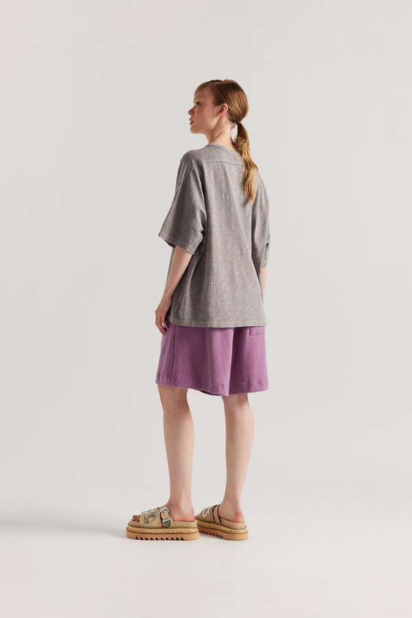 Back view of model in the Mauve Declare Tee in pigment-dyed slub cotton with relaxed boxy fit - highlighting the overlocked seam detailing. 