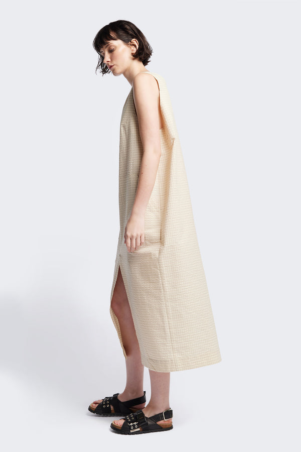 Side view of Long-length Forces Pinafore Dress in Ecru Beige Kloke designed soft seersucker premium cotton . Relaxed fit with a deep rounded neckline, front patch pockets, and a front split for ease of movement. Available in 5 sizes. 