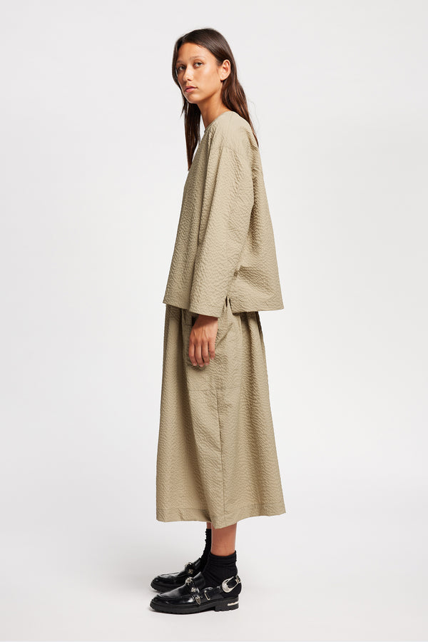 Side view of model wearing the Stone/beige Minor Skirt, an A-line midi skirt with an elastic waistband and soft gathered detailing, in lightweight Japanese seersucker.