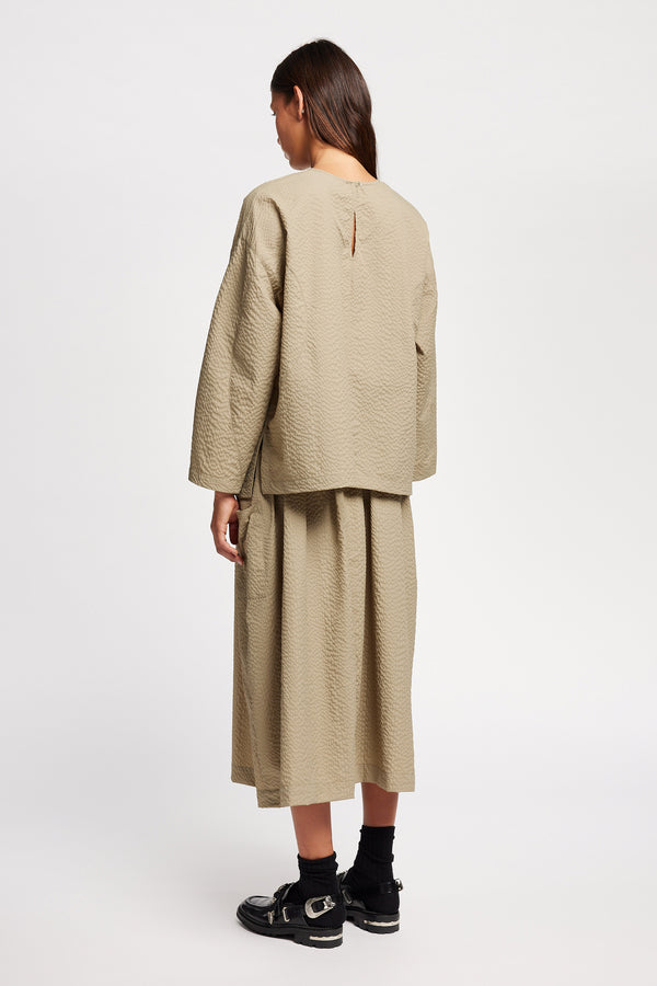 Back view of model wearing the Stone/beige Minor Skirt, an A-line midi skirt with an elastic waistband and soft gathered detailing, in lightweight Japanese seersucker.