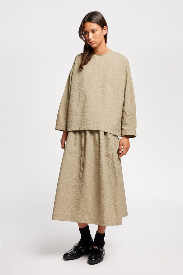 Front view of model wearing the Stone/beige Minor Skirt, an A-line midi skirt with an elastic waistband and soft gathered detailing, in lightweight Japanese seersucker.