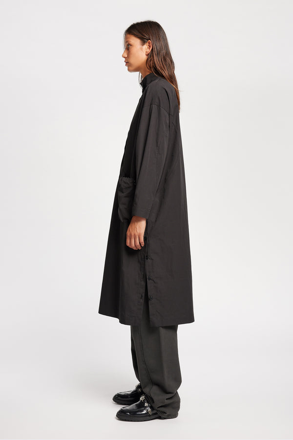 Side of Model wearing the Odeum Dress in black mid-weight cotton, featuring a high mock collar and large utility pockets.