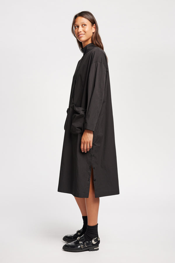Side of Model wearing the Odeum Dress in black mid-weight cotton, featuring a high mock collar and large utility pockets.
