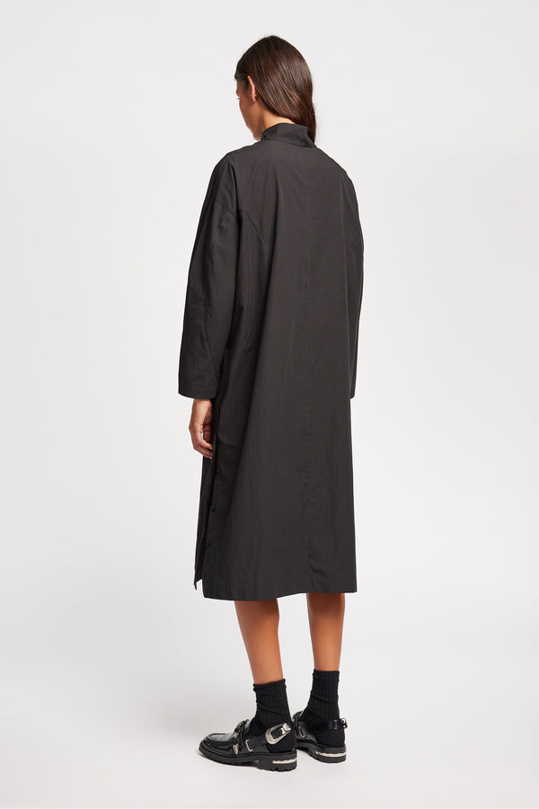 Back of Model wearing the Odeum Dress in black mid-weight cotton, featuring a high mock collar and large utility pockets.