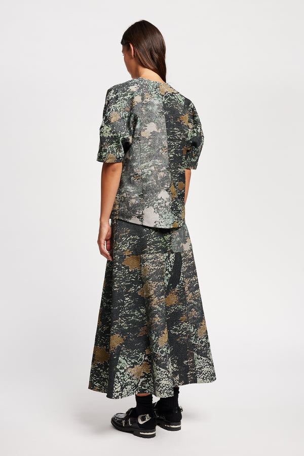 Back view model in the Optic Skirt a midi-length A-line skirt in textured jacquard green print fabric with a forest-inspired pattern, designed by Kloke