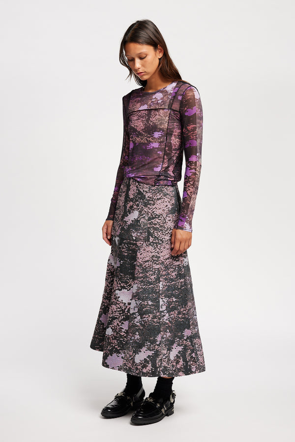 Front view model in the Optic Skirt a midi-length A-line skirt in textured jacquard purple print fabric with a forest-inspired pattern, designed by Kloke