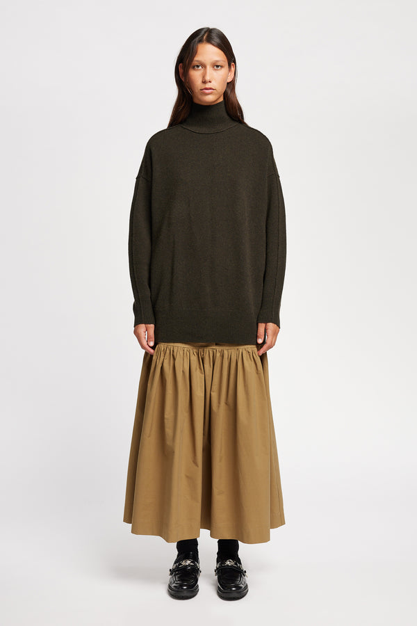 Front view of Model wearing Overflow long knit, high neck wool blend sweater with exposed seams in darkest green