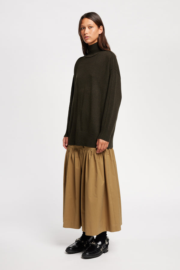 Front angle view of Model wearing Overflow long knit, high neck wool blend sweater with exposed seams in darkest green