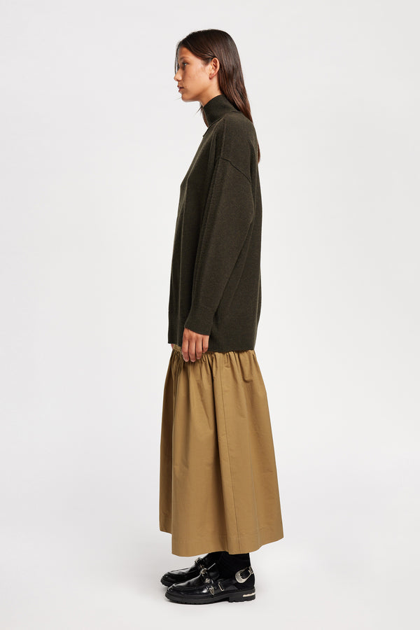 Side view of Model wearing Overflow long knit, high neck wool blend sweater with exposed seams in darkest green