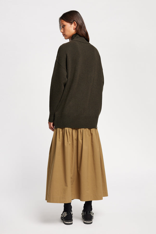 Back angle view of Model wearing Overflow long knit, high neck wool blend sweater with exposed seams in darkest green