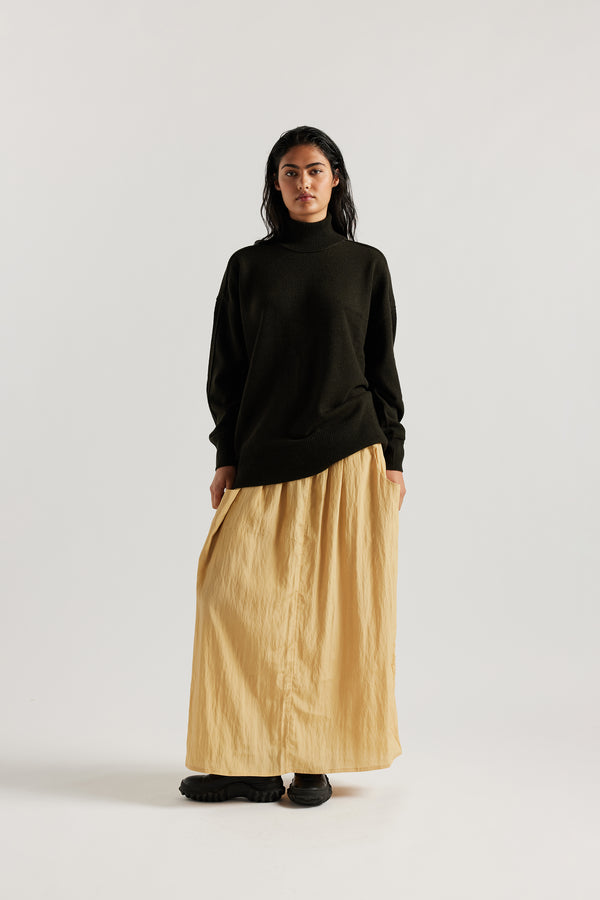 Front angle view of Model wearing Overflow long knit, high neck wool blend sweater with exposed seams in darkest green