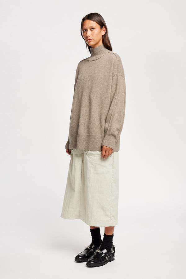 Front angle view of Model wearing Overflow long knit, high neck wool blend sweater with exposed seams in taupe