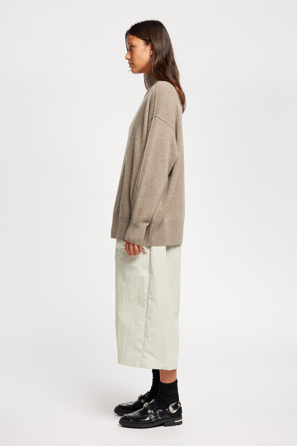 Side view of Model wearing Overflow long knit, high neck wool blend sweater with exposed seams in taupe