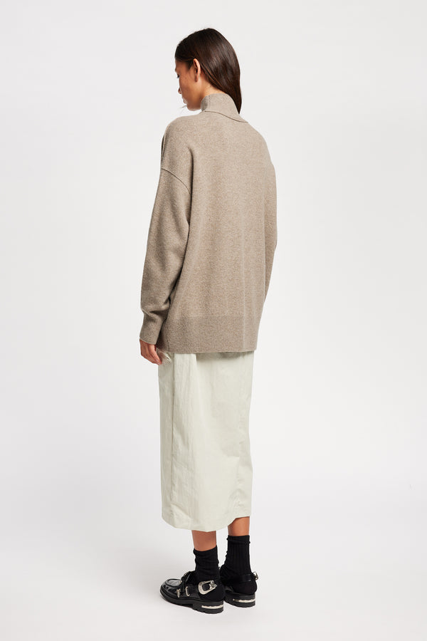 Back angle view of Model wearing Overflow long knit, high neck wool blend sweater with exposed seams in taupe